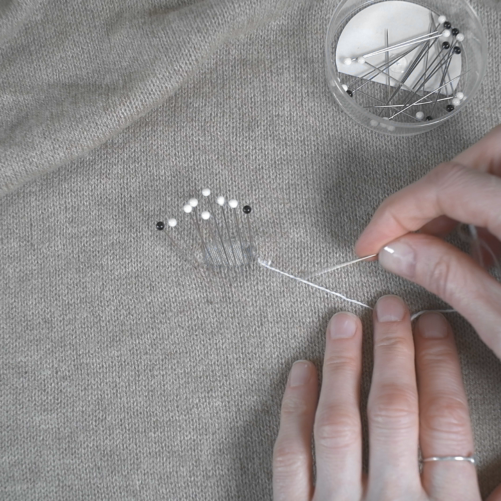 Person sewing on cashmere with a small container of pins nearby.